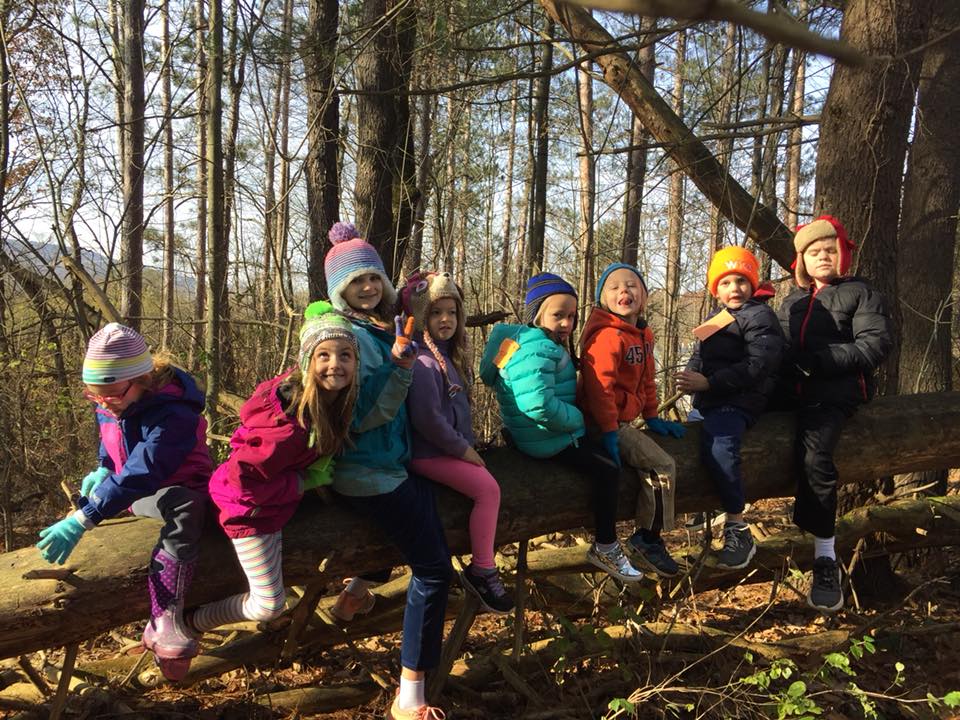 Young children sitting on a log in the woods, some making funny faces